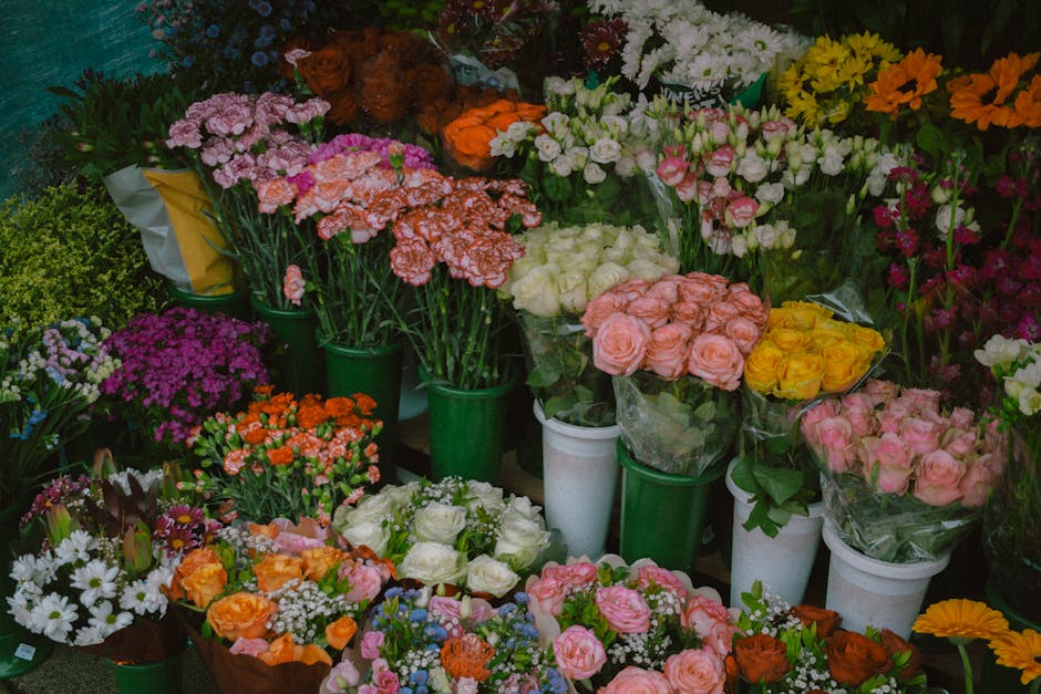 A vibrant display of fresh floral arrangements featuring pink, orange, white, yellow, and purple flowers, including roses, carnations, daisies, and chrysanthemums, arranged in green and white vases an