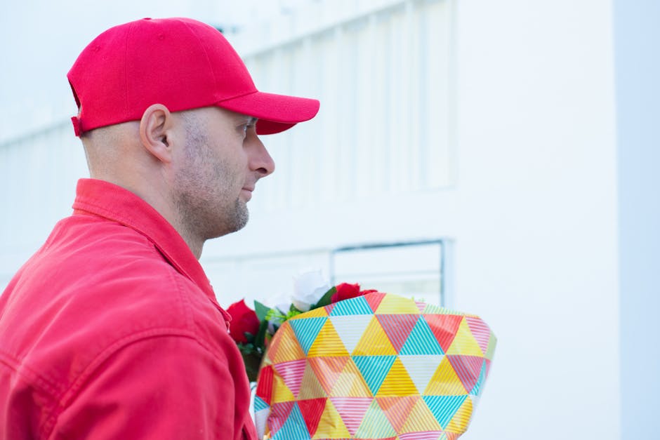 A man dressed in a red jacket and matching red cap holding a bouquet of fresh flowers wrapped in colorful geometric-patterned paper, with a focus on the vibrant reds, whites, and greens of the floral 
