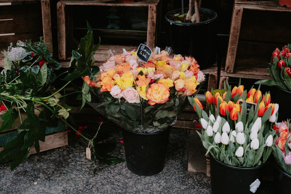 A black flowerpot filled with a bouquet of fresh, multicolored roses in shades of yellow, pink, and peach, arranged with dark green foliage. The bouquet has a lush, rounded shape and is wrapped in cle