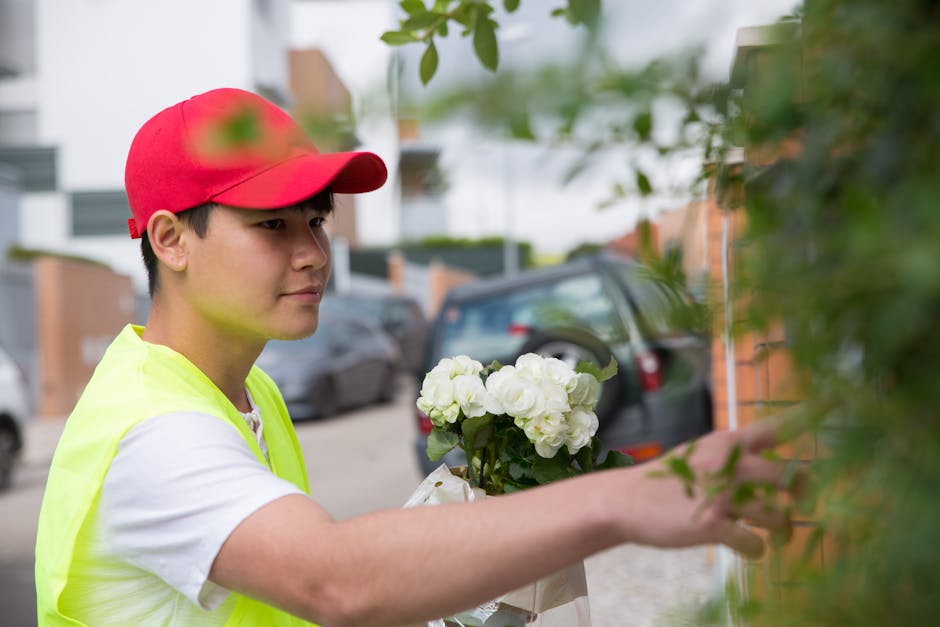 A young male flower delivery courier wearing a red cap and a neon yellow vest is outdoors, holding a bouquet of fresh white roses and hydrangeas wrapped in white paper. He is reaching out to place the