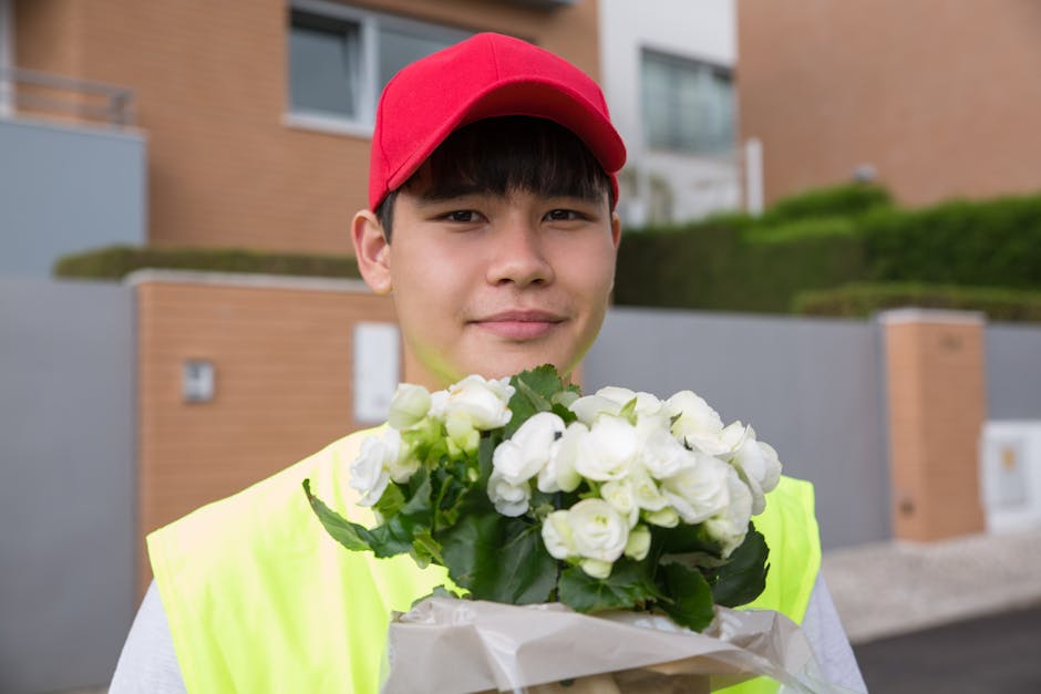 A young delivery person wearing a bright yellow high-visibility vest and a red cap is holding a bouquet of fresh white roses wrapped in white paper. The flowers are well-arranged with lush green leave